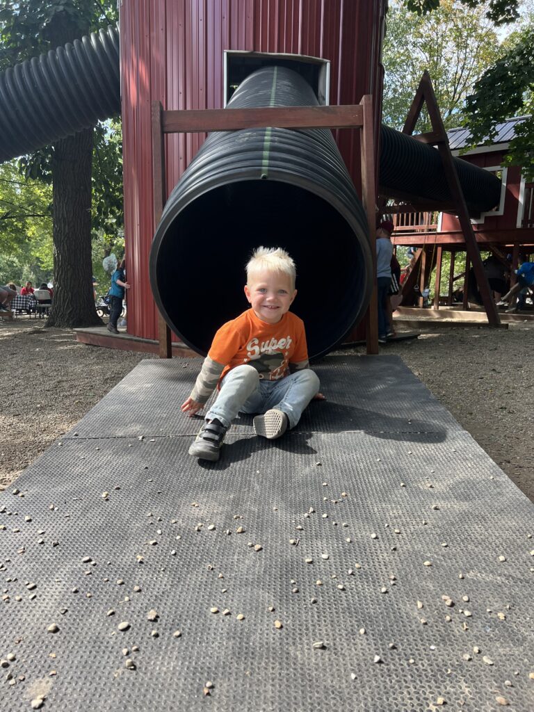 Smiling young blond boy in an orange 'Super' shirt and jeans sits at the exit of a large black corrugated tube slide attached to a red wooden play structure in a tree-shaded playground.