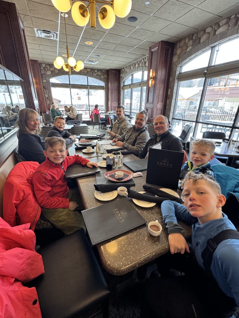 A family group of adults and children seated around a long restaurant table inside a lodge-style dining room, with menus, coffee cups and winter jackets on the table and large windows showing a snowy ski-area scene outside.
