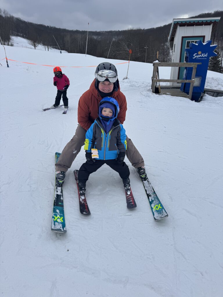 Adult in a red jacket and black helmet crouches behind a small child in a blue jacket and helmet as they both balance on skis on a snowy beginner slope, with another child in a pink jacket behind them and a small SunKid booth to the right.