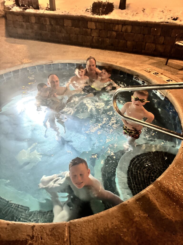 A group of adults and children relaxing in a steaming round outdoor hot tub at night, one boy wearing goggles by the metal handrail and snow visible on the stone deck behind them.