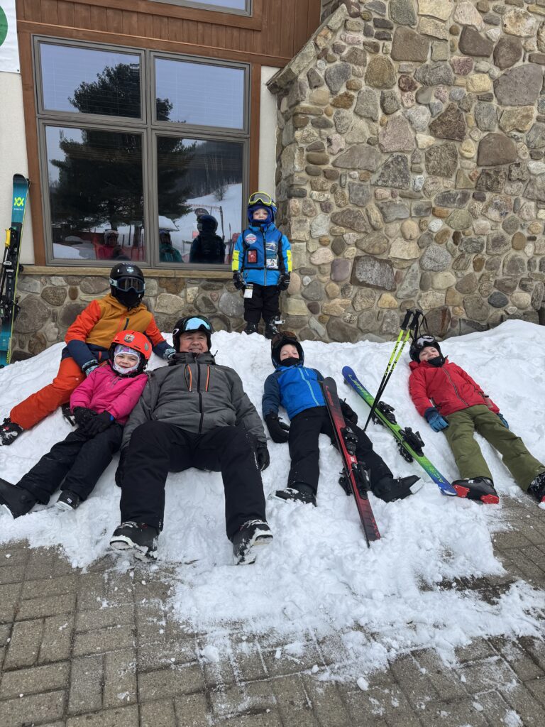 A group of skiers in helmets and colorful winter gear — one child standing in a blue jacket with yellow goggles and five people reclining on a snowbank with skis and poles — in front of a stone lodge wall and a large window reflecting trees.