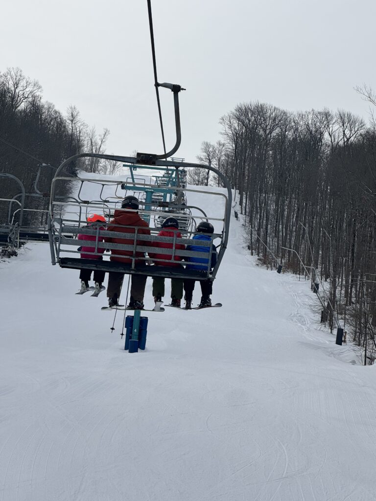 Five people wearing helmets and colorful winter jackets sit on a chairlift with skis and snowboards dangling as it ascends a groomed snowy slope lined by leafless trees under an overcast sky.