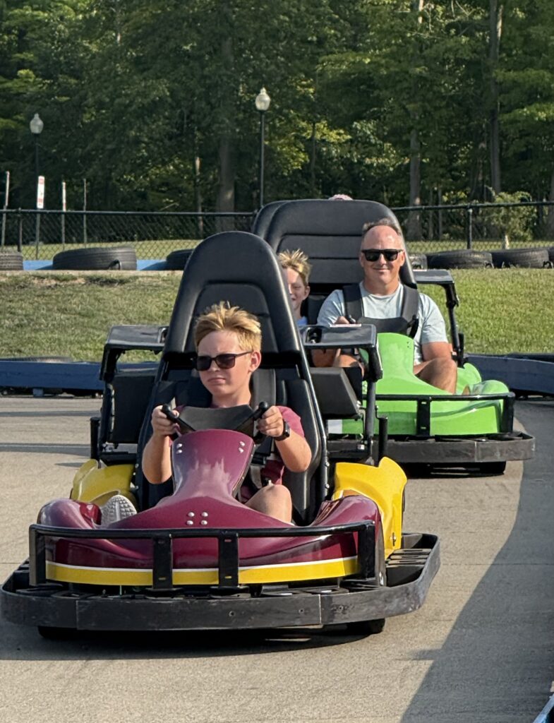 Teenage boy wearing sunglasses drives a maroon-and-yellow go-kart with a focused expression, followed by a smiling man in sunglasses in a green go-kart and another child between them on an outdoor track with trees in the background.