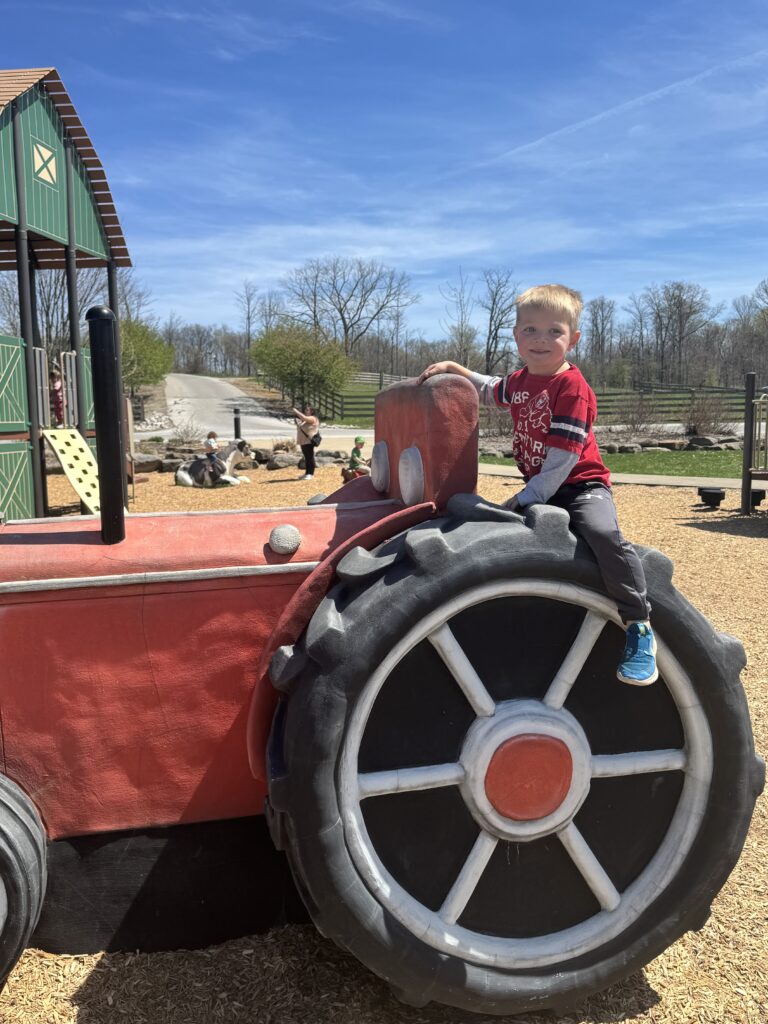 A young boy in a red shirt and blue sneakers sits smiling on the large black-and-red wheel of a play tractor at an outdoor playground with wood-chip ground, a green play barn, and trees under a clear blue sky.