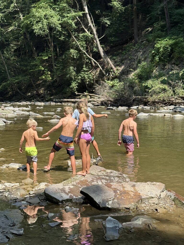 Five children in swimsuits wade and balance on rocks in a shallow, tree-lined stream with sunlight dappled on the water and rocky shore.
