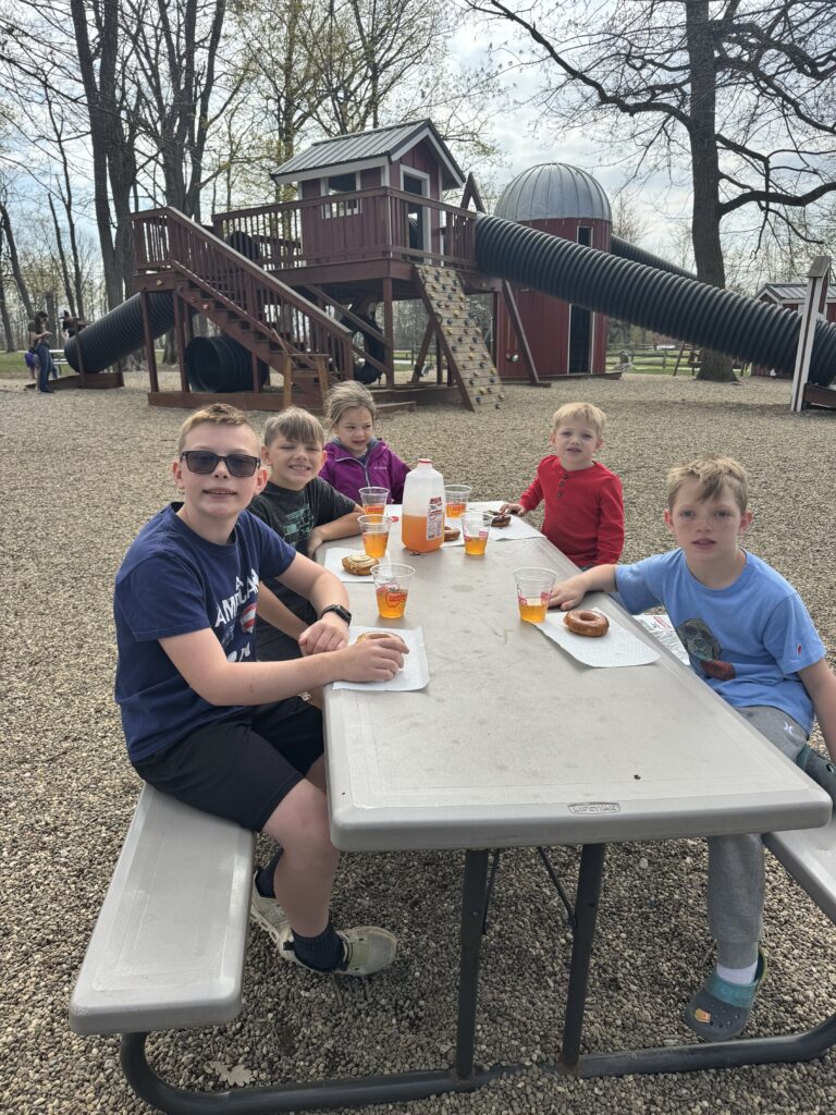 Five children sit around a picnic table eating donuts and drinking an orange beverage at a gravel-surfaced playground in front of a wooden play structure with large corrugated tube slides and a climbing wall, with bare trees and an overcast sky behind them.