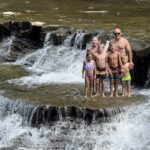 An adult man wearing sunglasses and six children in colorful swimsuits standing together on a flat rock ledge while water cascades over the surrounding rocks into a shallow waterfall below.