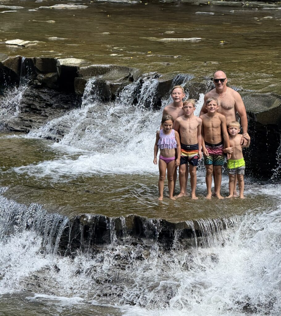 A shirtless man wearing sunglasses stands with five children in colorful swimsuits on a flat rock ledge while water cascades around them over a multi-tiered waterfall.