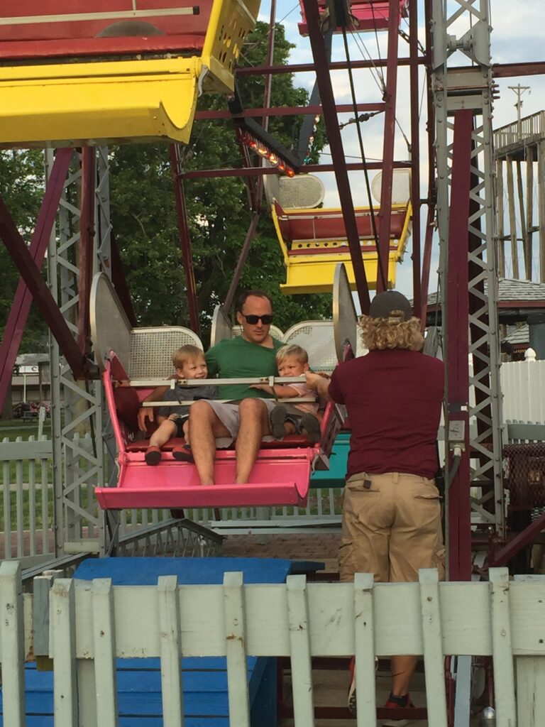 Man wearing sunglasses and two young boys seated in a pink Ferris wheel car as an attendant secures the safety bar at a small amusement ride with yellow gondolas and a white picket fence.