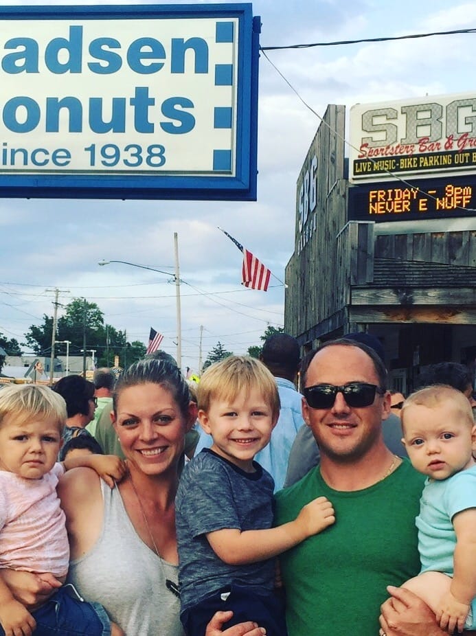 A smiling family of two adults and three young children posing outdoors in front of a donut shop sign and a wooden storefront with American flags and a marquee sign, father wearing sunglasses and mother holding a toddler.