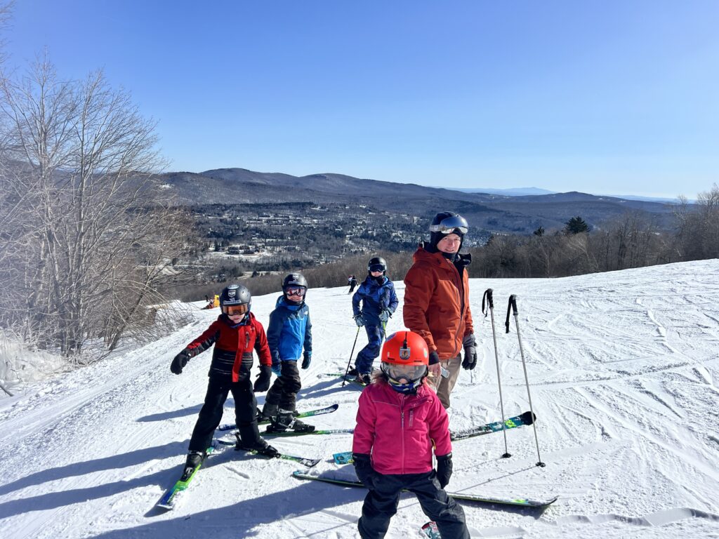Group of four skiers — three children in colorful jackets and helmets (front child in pink with a red helmet, two others in red and blue) and an adult in an orange jacket — standing on a snowy ski slope with skis and poles, overlooking a valley and distant tree-covered mountains under a clear blue sky.