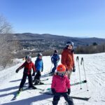 Group of four skiers — three children in colorful jackets and helmets (front child in pink with a red helmet, two others in red and blue) and an adult in an orange jacket — standing on a snowy ski slope with skis and poles, overlooking a valley and distant tree-covered mountains under a clear blue sky.