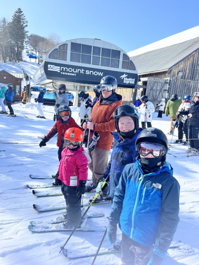 Four children in colorful ski jackets and helmets with an adult in an orange jacket stand on skis in front of the Mount Snow “Bluebird Express” lift on a sunny snowy day, with other skiers and a wooden lodge in the background.