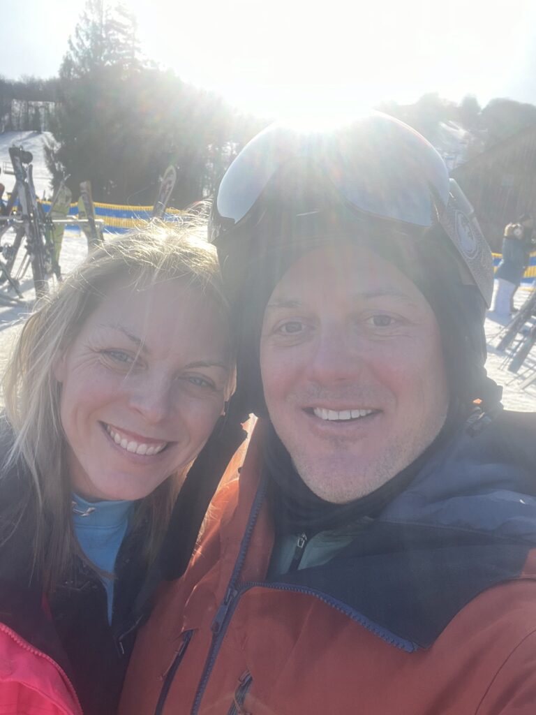 Close-up selfie of a smiling man in an orange ski jacket and helmet with goggles and a woman leaning in beside him, bright sun glare and snowy ski slopes with parked skis visible behind them.