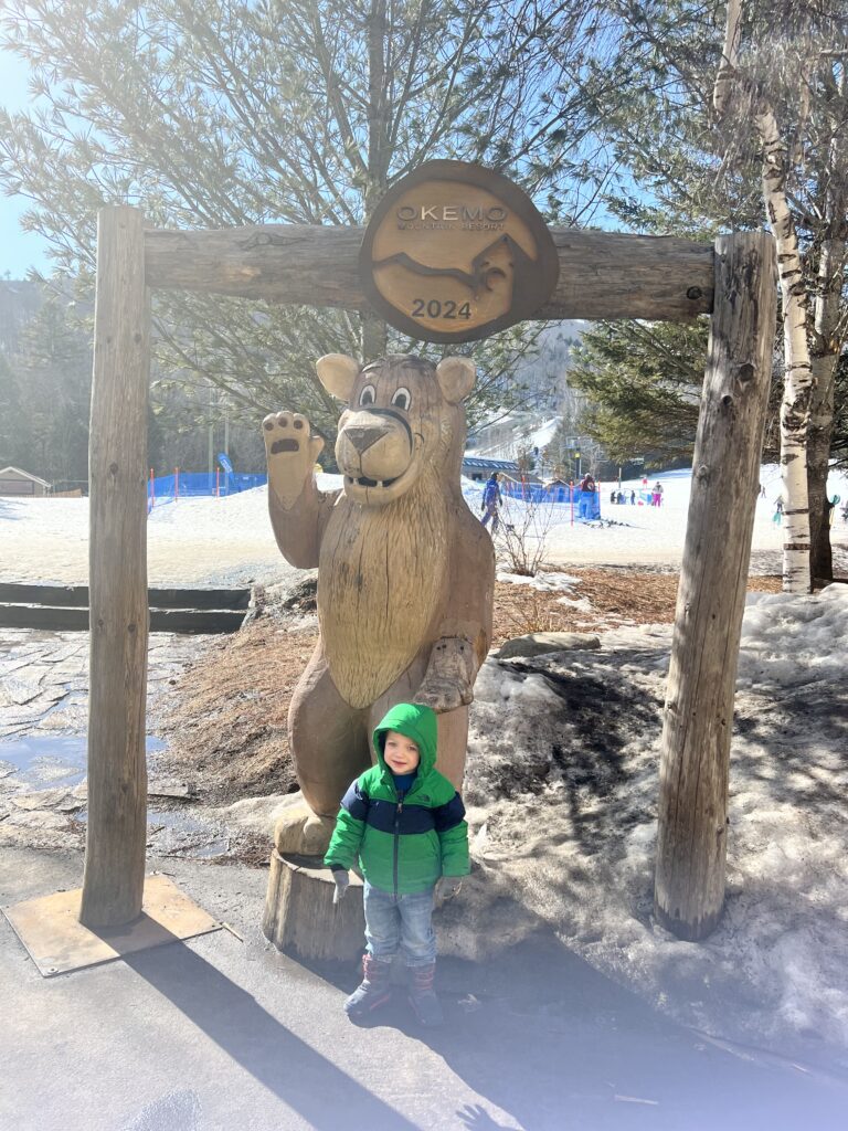 Young child in a green hooded jacket and snow boots stands smiling in front of a carved wooden bear under a log arch marked “Okemo Mountain Resort 2024,” with snow-covered ski slopes and trees in the background.