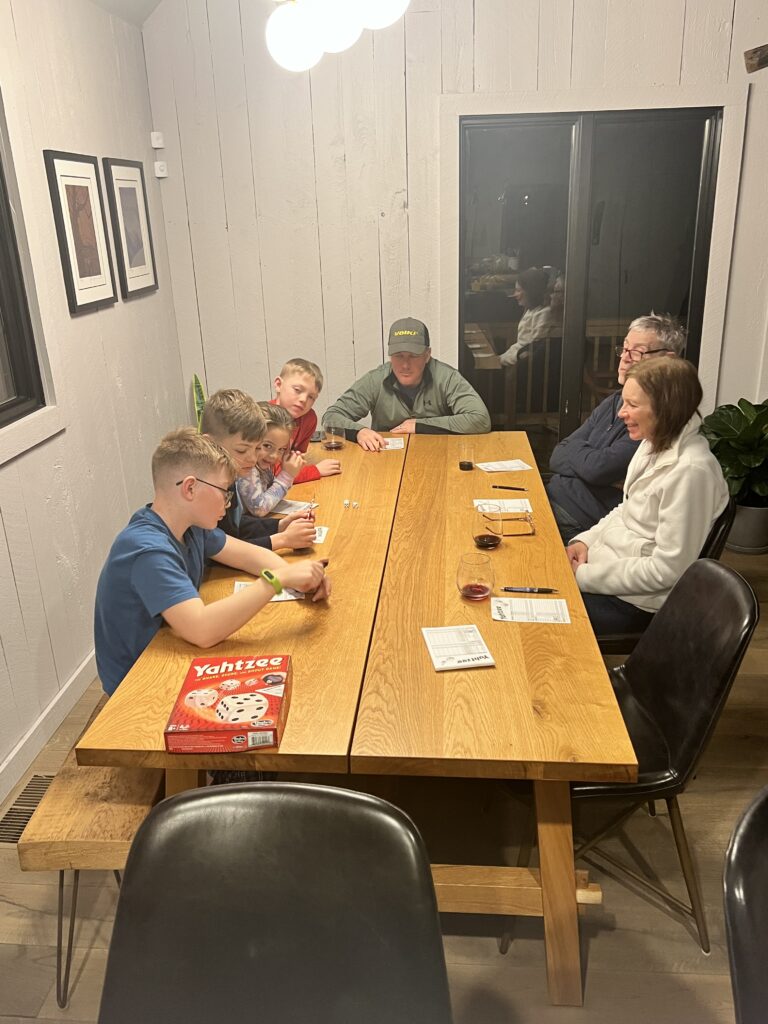 Group of family members seated around a long oak dining table playing Yahtzee — four children on the left rolling dice and filling scorecards, two adults on the right watching and sipping wine, with the red Yahtzee box visible near the table's end.