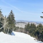 Snow-covered ski slope descending between evergreen trees with snow guns on the left and hazy blue mountain ridgelines under a clear blue sky.