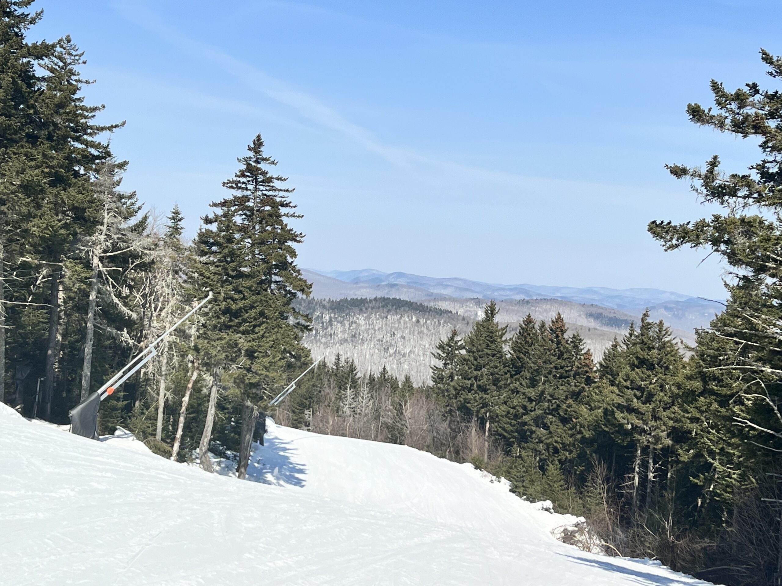 Snow-covered ski slope descending between evergreen trees with snow guns on the left and hazy blue mountain ridgelines under a clear blue sky.