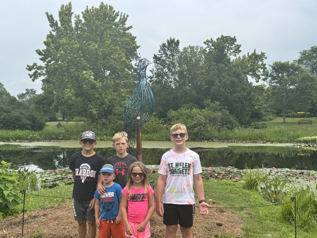 Five children standing on a grassy path in front of a lily-pad covered pond and a tall blue wire peacock sculpture, with dense green trees and a park bench in the background.