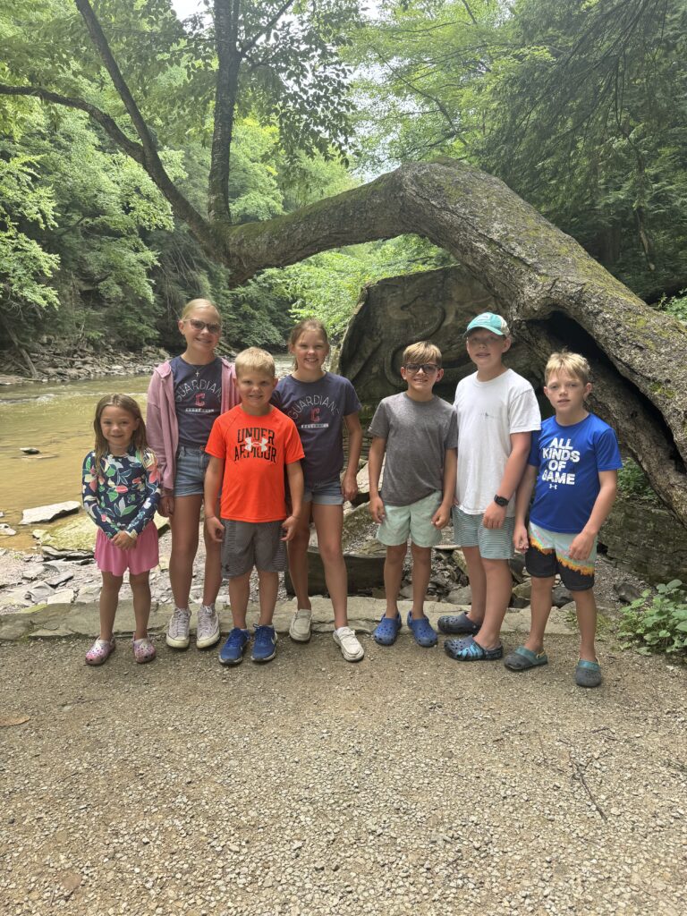 Eight children and preteens stand shoulder-to-shoulder on a gravel path by a shallow river, smiling in casual summer clothes and sneakers/Crocs, with a large mossy curved tree arching over them and dense green forest behind.