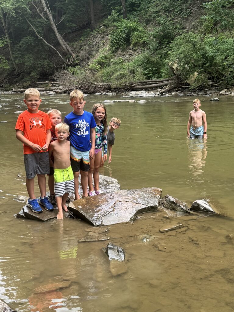 Seven children play in a shallow, muddy river with a wooded, rocky bank behind them — six stand together on flat rocks near the near shore wearing swimsuits and colorful shirts while one boy wades alone farther out.