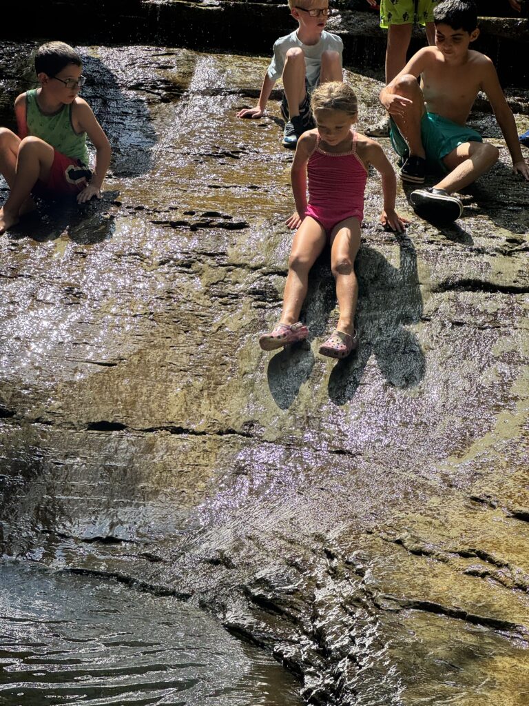 Several children in swimsuits sit and slide on a sunlit wet rock slope above a shallow pool, the central girl in a pink one-piece with her legs extended while others watch nearby.
