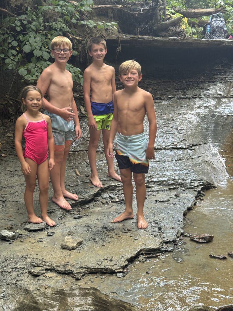 Four children in swimsuits standing barefoot on a wet, layered rock ledge beside a shallow creek in a wooded area, one boy wearing glasses and a backpack resting on a log in the background.