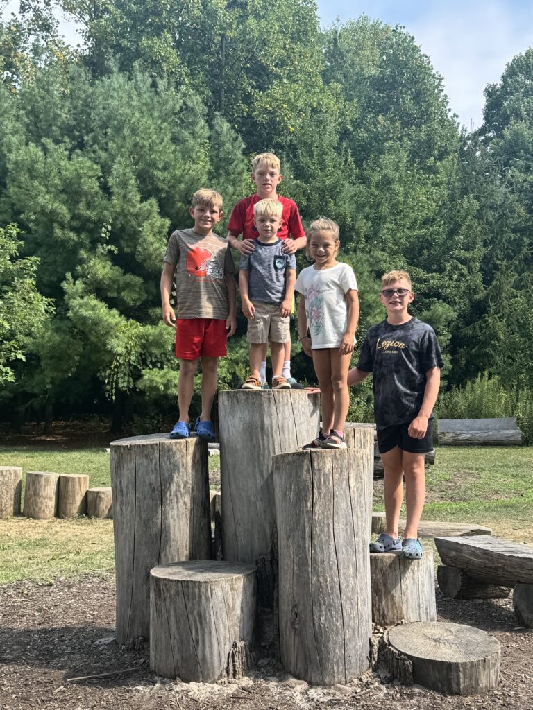 Five children pose on a cluster of tall cut tree stumps in a grassy park with dense green trees behind them, wearing casual summer clothes (red shirt center back; gray shirt with orange helmet graphic and red shorts left; small striped shirt and khaki shorts front center; white T-shirt and shorts right-center; dark shirt and Crocs far right).