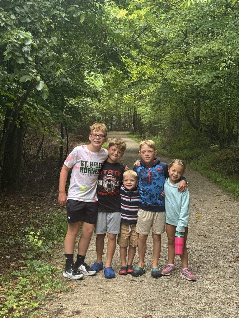 Five smiling children stand close together with arms around each other on a shaded gravel forest trail under a green tree canopy, wearing casual summer clothes and sneakers or Crocs, one girl holding a pink water bottle.