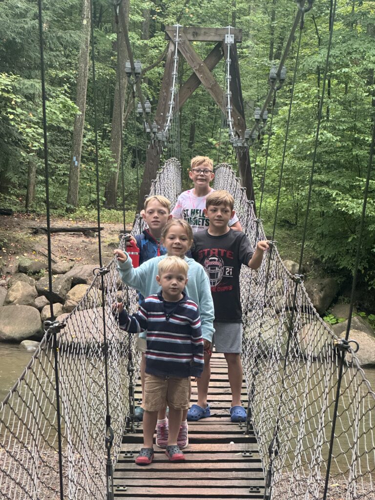 Five children stand in a line on a narrow wooden-plank suspension bridge with rope-net sides over a shallow stream, framed by rocks and dense green forest.