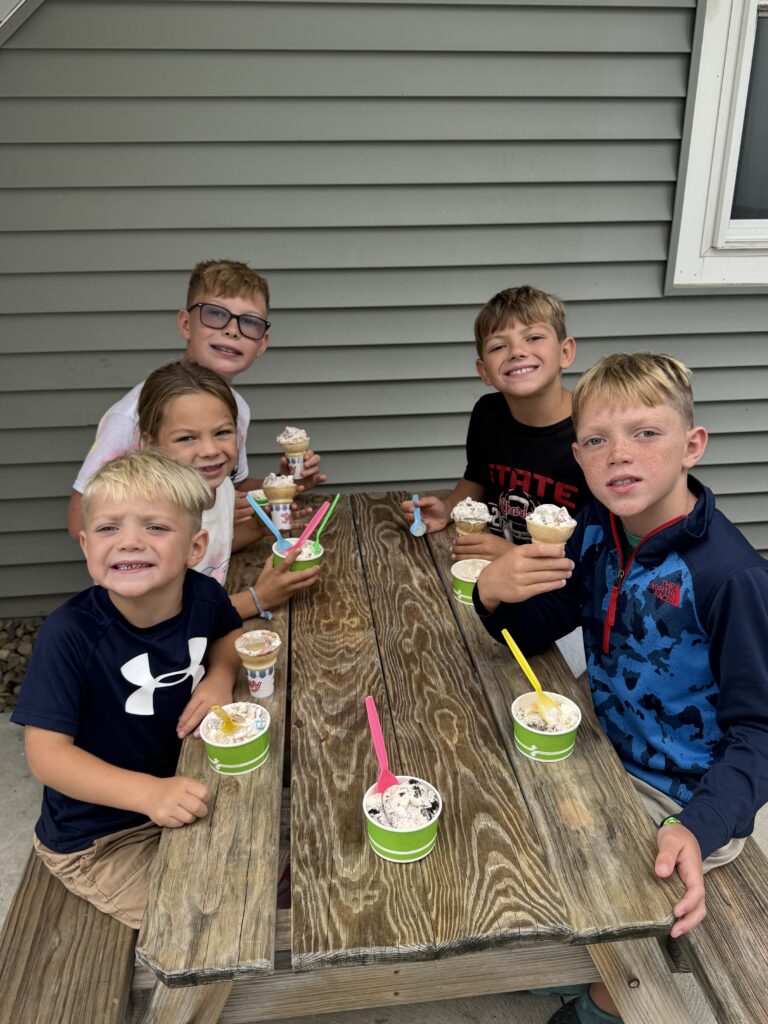 Six smiling children seated around a weathered wooden picnic table outdoors, each holding small cups or cones of ice cream with colorful spoons, set against gray horizontal house siding and a window.