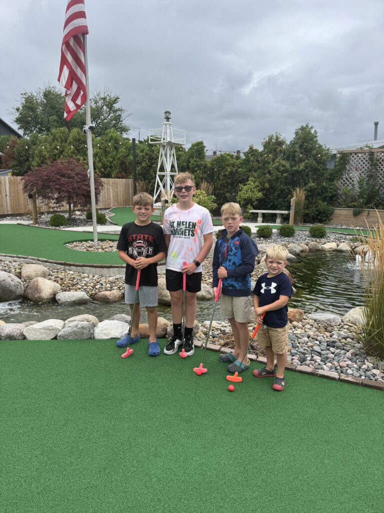 Four boys standing on a miniature golf green holding colorful plastic putters, with a rocky pond, decorative white windmill, American flag and cloudy sky in the background.