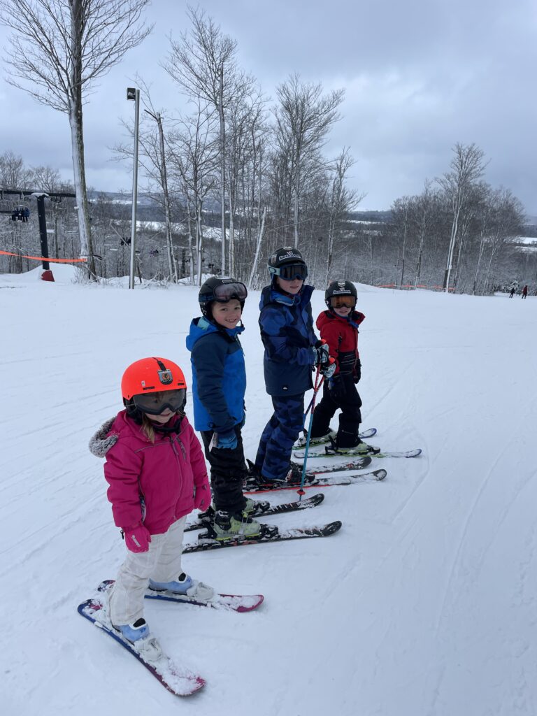 Four children in colorful ski gear—front child in a pink jacket and bright orange helmet, two children in blue jackets and helmets, and a child at the back in red—standing on skis on a snow-covered slope with bare trees and an overcast sky.