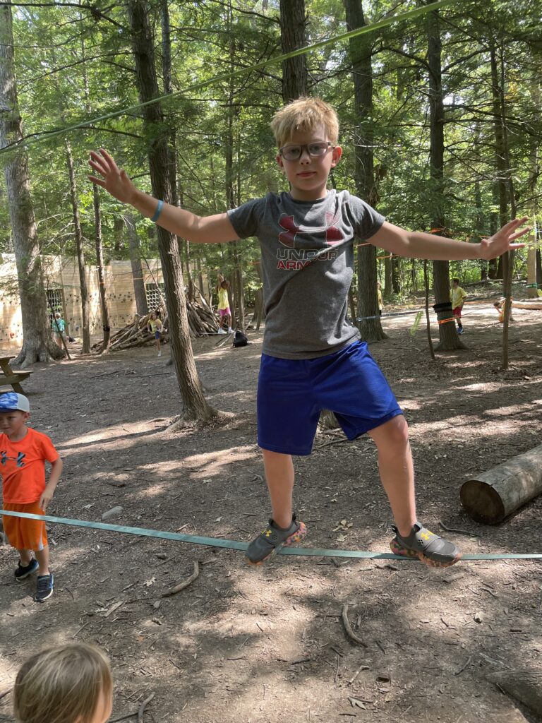 Young boy wearing glasses, a gray Under Armour t-shirt and blue shorts balances with arms outstretched on a slackline in a shaded forest activity area, with other children and a climbing wall visible in the background.