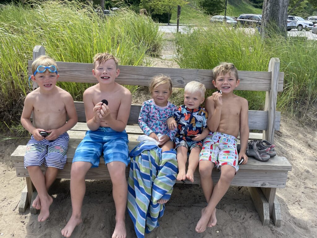Five young children in swimwear sit side-by-side on a wooden bench at a sandy beach with tall grasses behind them; three boys hold cookies, a girl in a floral long-sleeve swimsuit wrapped in a striped towel sits in the center, and a toddler in a colorful rash guard sits next to her.