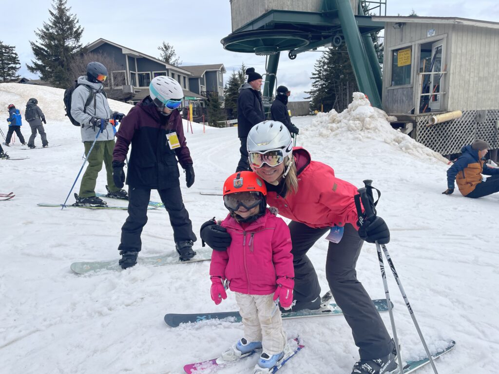 Woman in a bright pink ski jacket and white helmet crouches beside a young child in a red helmet and pink jacket, both on skis near a chairlift terminal with other skiers and snowy lodge buildings in the background.