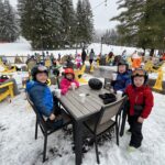 Four children in ski helmets and winter jackets sitting and standing around a wooden patio table holding cups of hot drink at a snowy ski-lodge terrace, with yellow Adirondack chairs, other skiers, and snow-covered evergreen trees and ski slopes in the background.