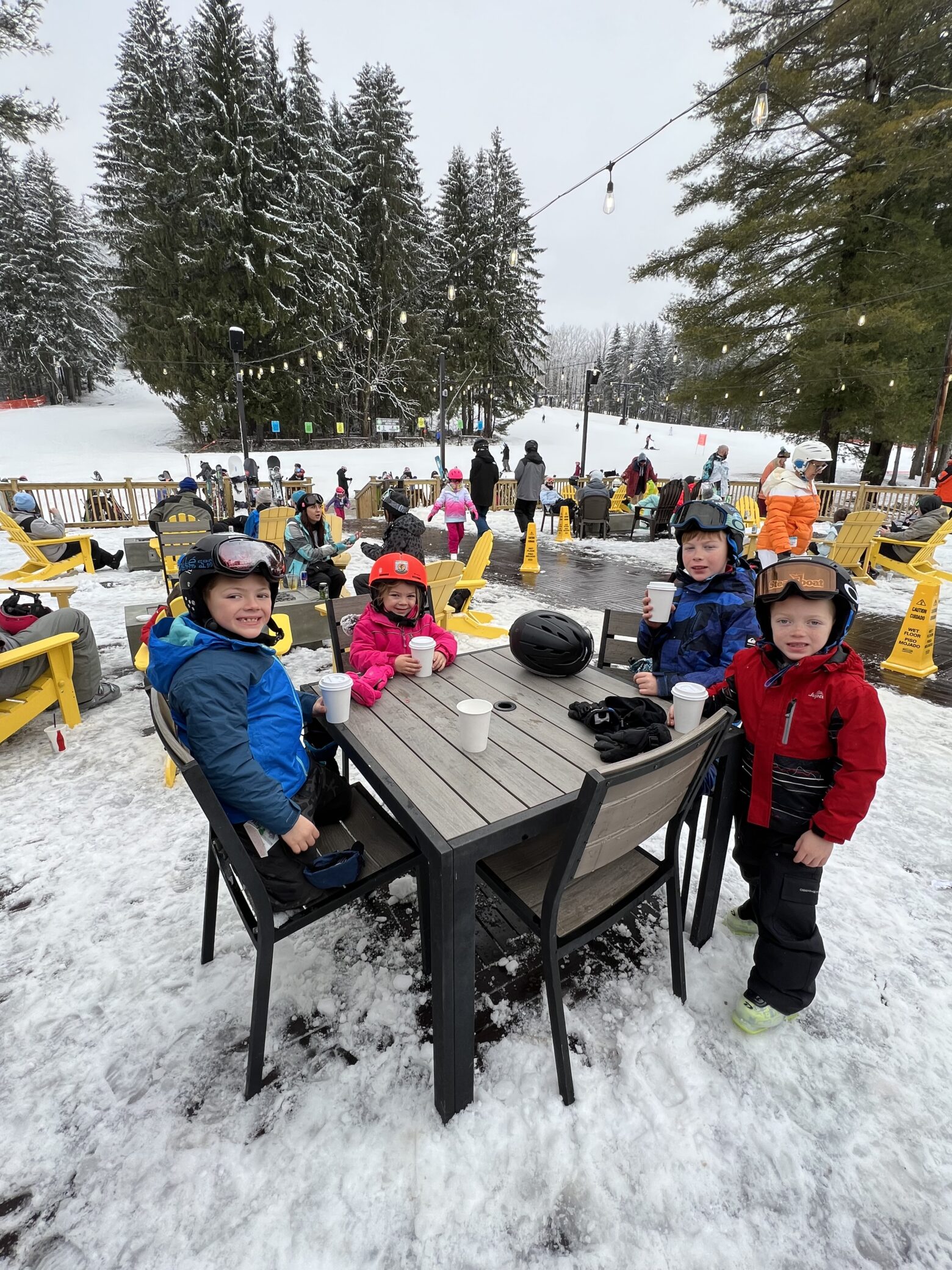 Four children in ski helmets and winter jackets sitting and standing around a wooden patio table holding cups of hot drink at a snowy ski-lodge terrace, with yellow Adirondack chairs, other skiers, and snow-covered evergreen trees and ski slopes in the background.