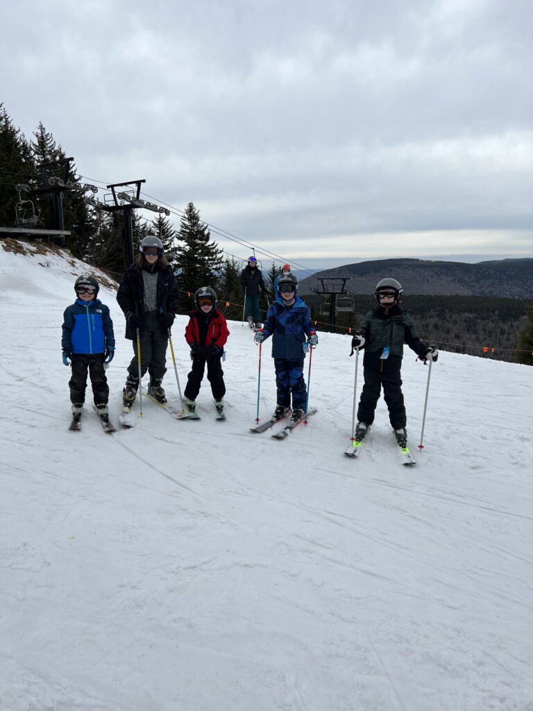 Five skiers, including four children and one taller adult, stand on a snowy slope in helmets and colorful ski gear near a chairlift, with forested hills and an overcast sky behind them.