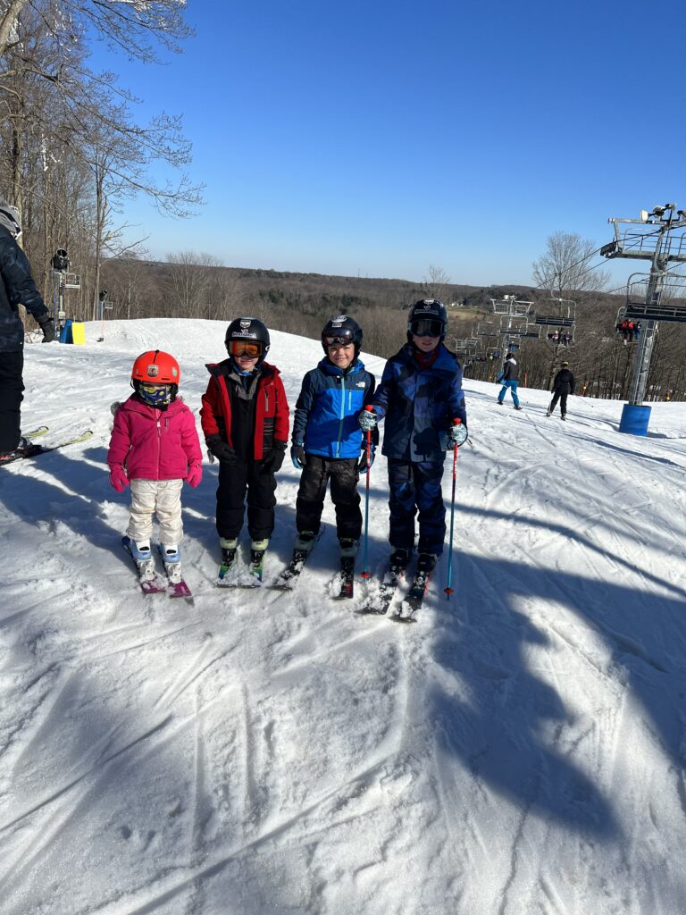 Four children in helmets and ski gear—a small girl in pink and three boys in red and blue jackets—stand on a snowy ski slope with skis and poles, a chairlift to the right and bare trees under a clear blue sky.
