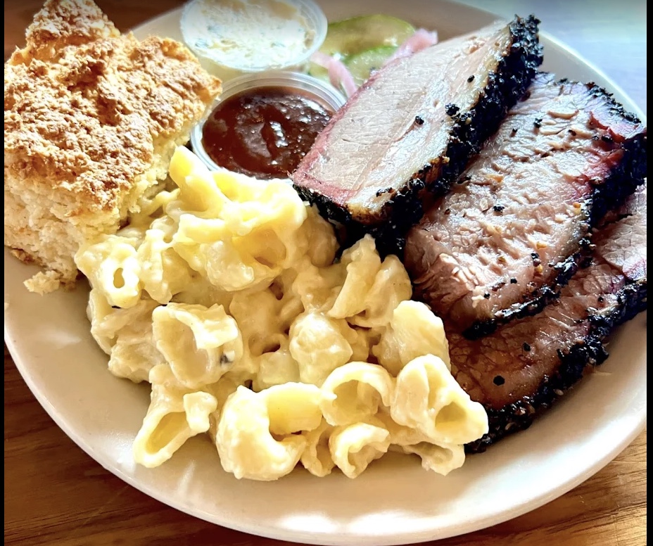 Plate of barbecue featuring several thick slices of smoked brisket with a dark peppery bark and pink smoke ring, creamy shell macaroni and cheese, a flaky biscuit, and small containers of barbecue sauce and pickles.