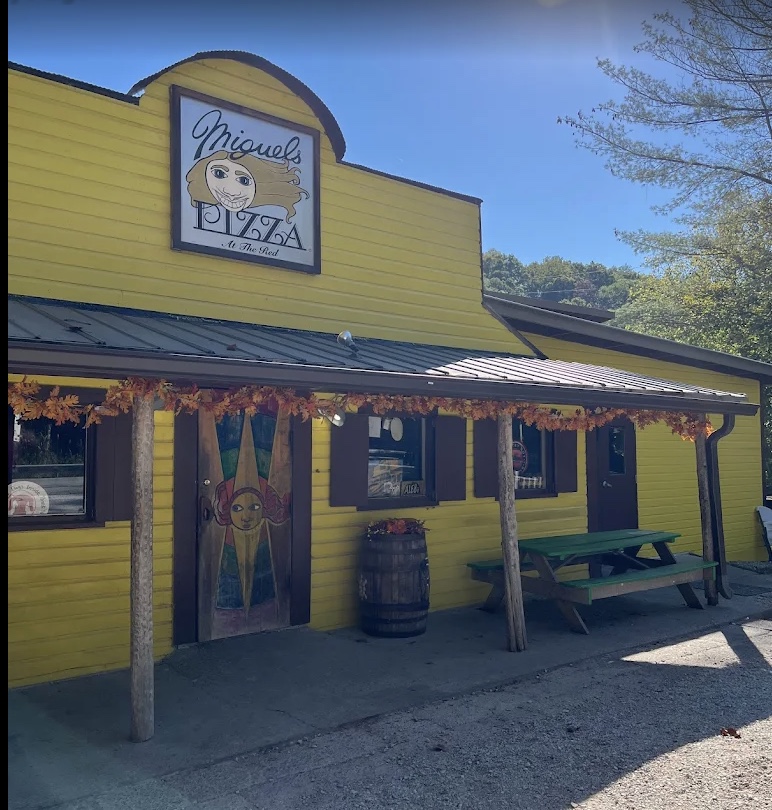 Bright yellow wooden pizza shop with a 'Miguel's Pizza' sign, a painted door featuring a sun face, a porch trimmed with autumn garlands on log posts, a wooden barrel planter and a green picnic table outside.
